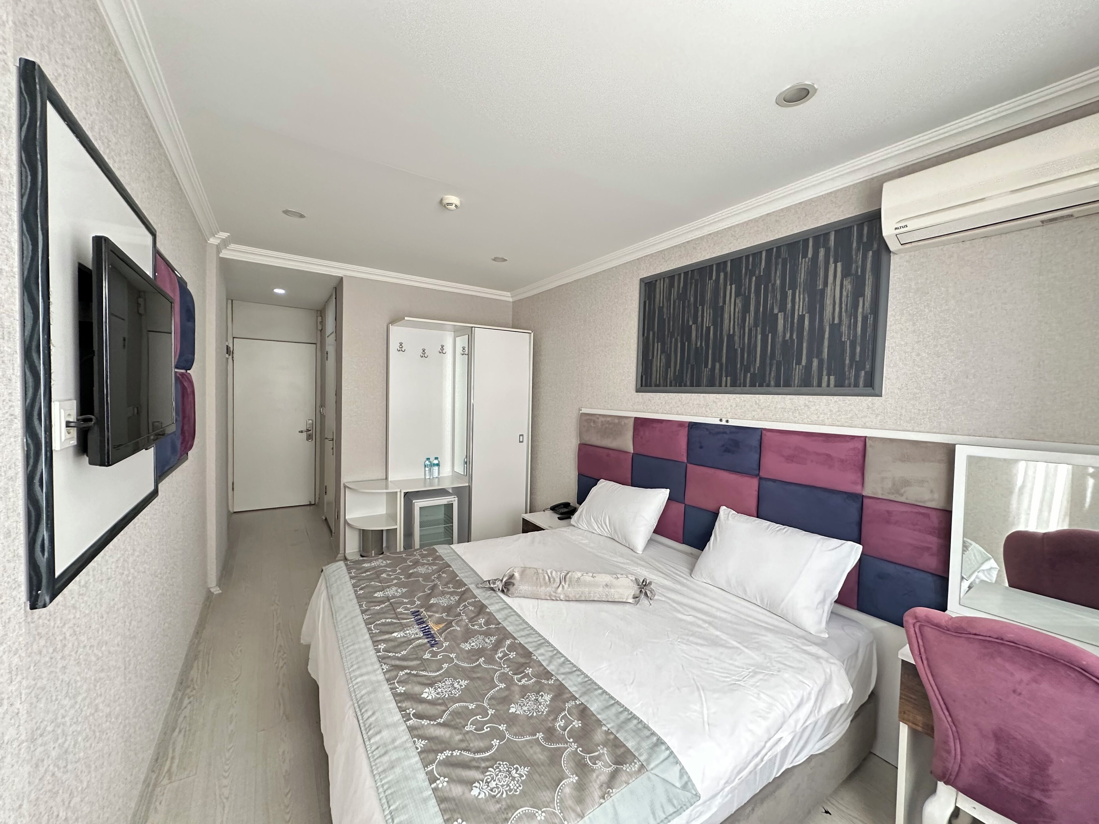 A hotel room with a bed, bedside table, mirror, wardrobe, and an air conditioning unit.