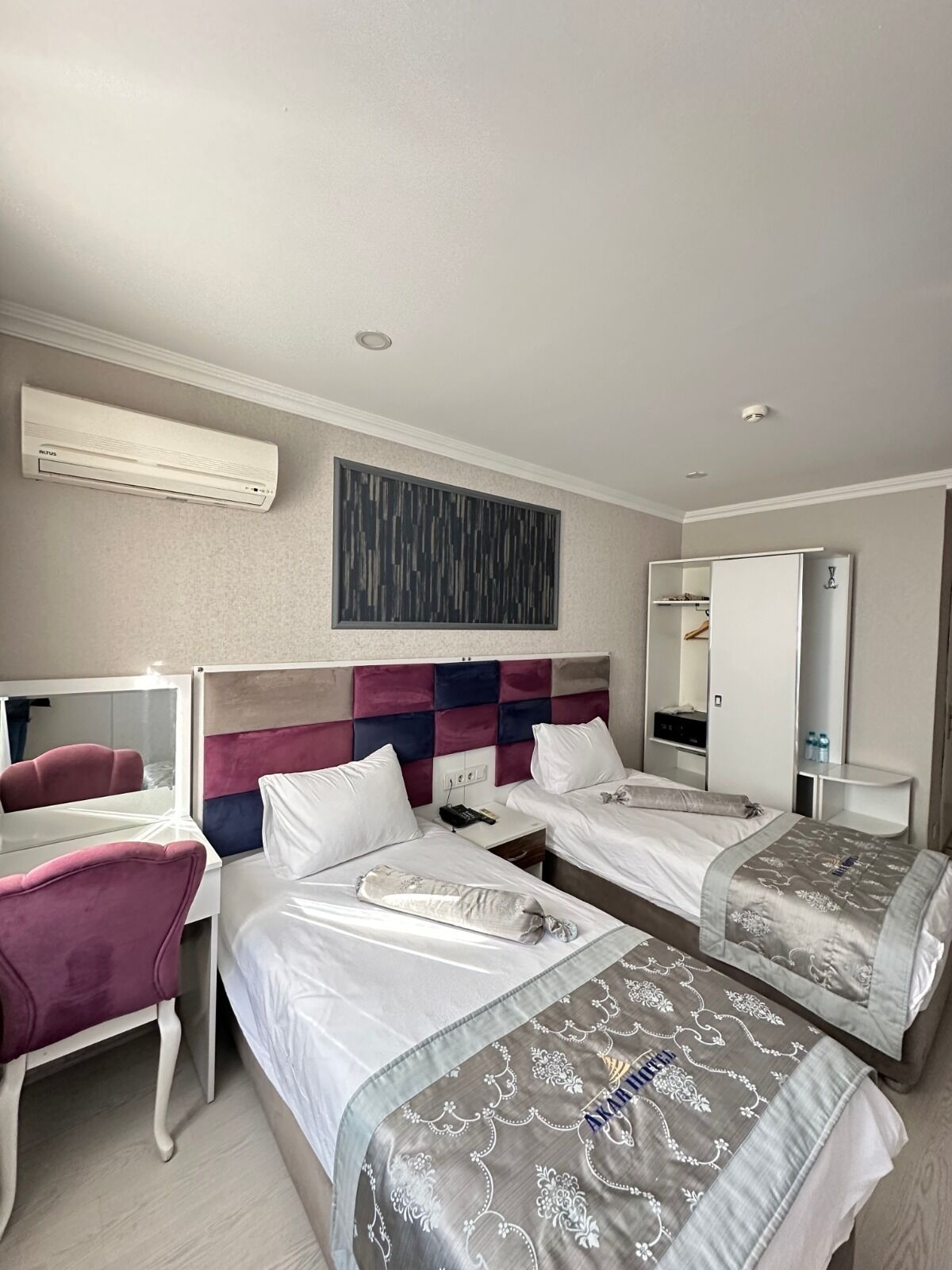 A hotel room with two beds, a vanity with a mirror, a wardrobe, and an air conditioning unit.