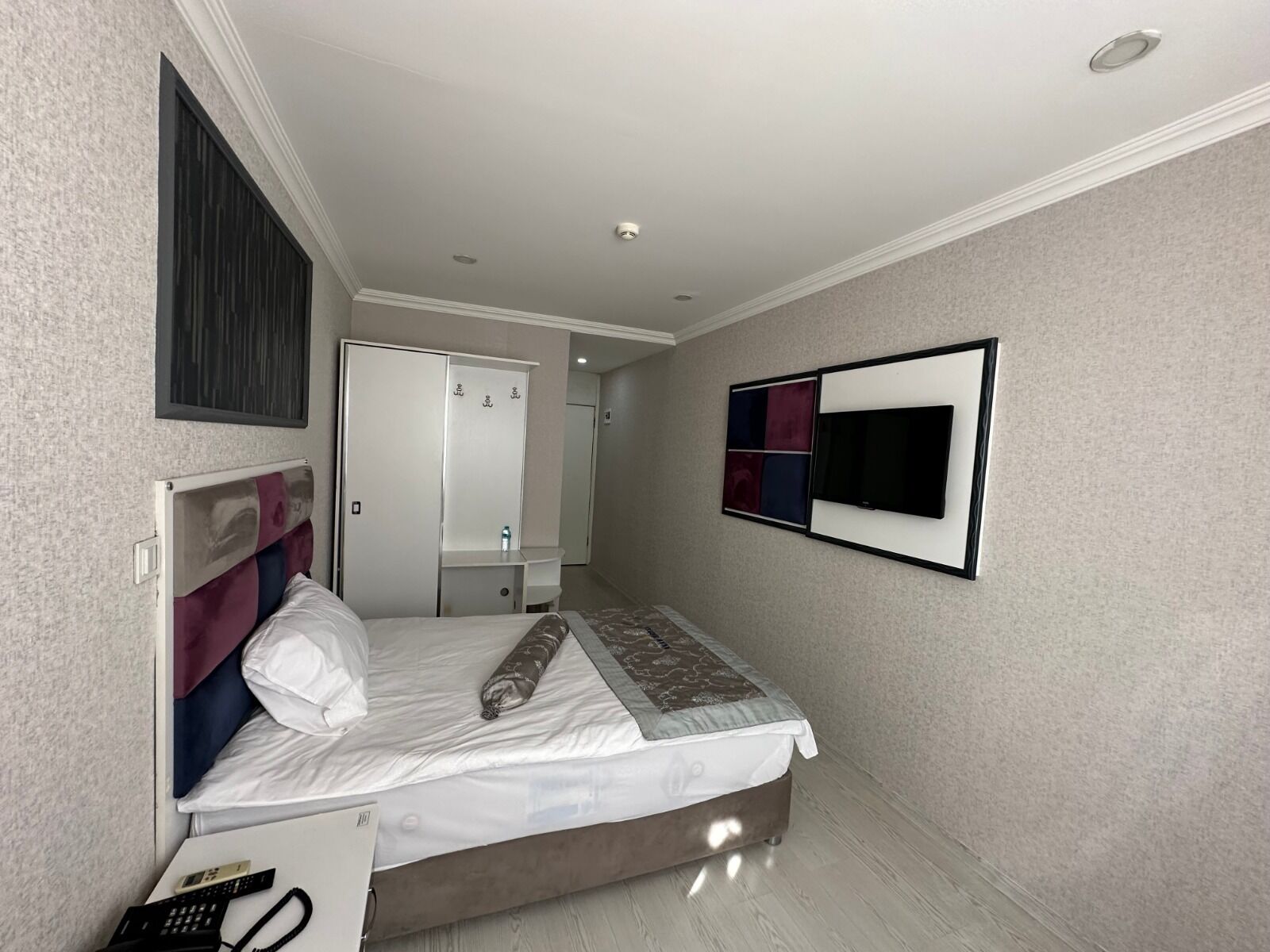 A hotel room with a bed, a bedside table with a phone, a mirror, a wardrobe, and a TV mounted on the wall.