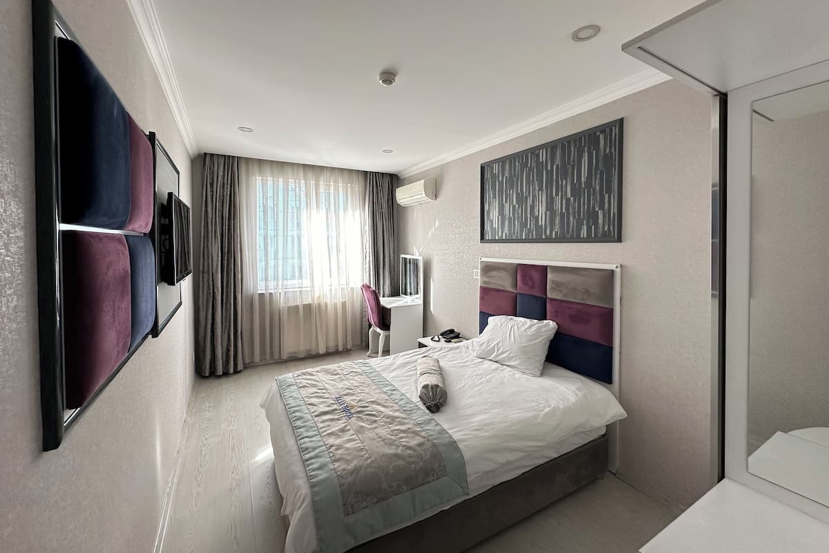 Una habitación de hotel moderna con una cama grande, un televisor de pantalla plana en la pared, un escritorio con una silla y una ventana con cortinas translúcidas.