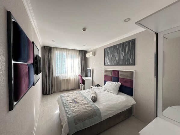 A modern hotel room with a large bed, a flat-screen TV mounted on the wall, a desk with a chair, and a window with sheer curtains. - Akar otel (Elazig)
