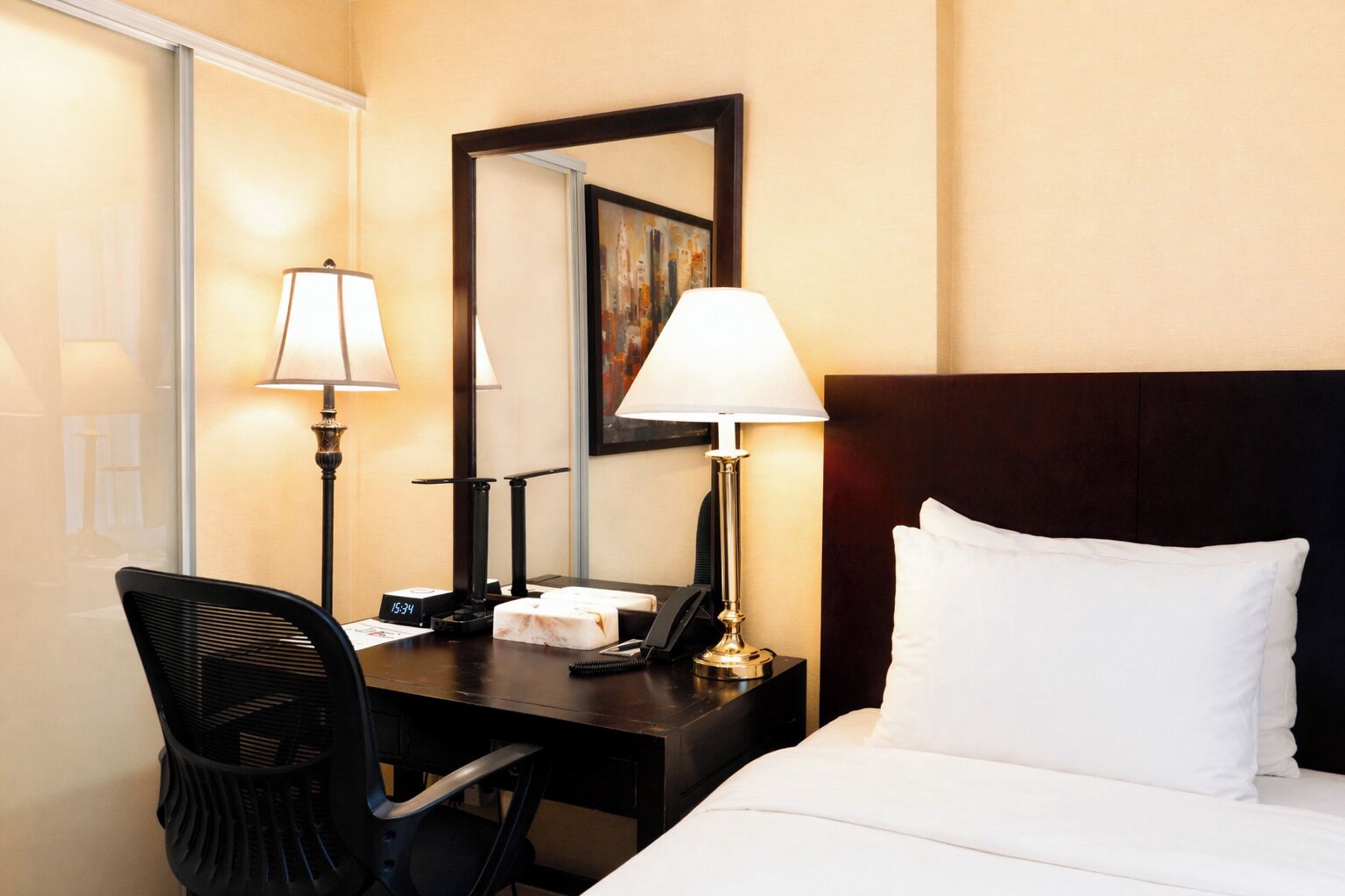 Habitació d'hotel amb un llit, un escriptori, una cadira, una làmpada, un rellotge i un mirall.