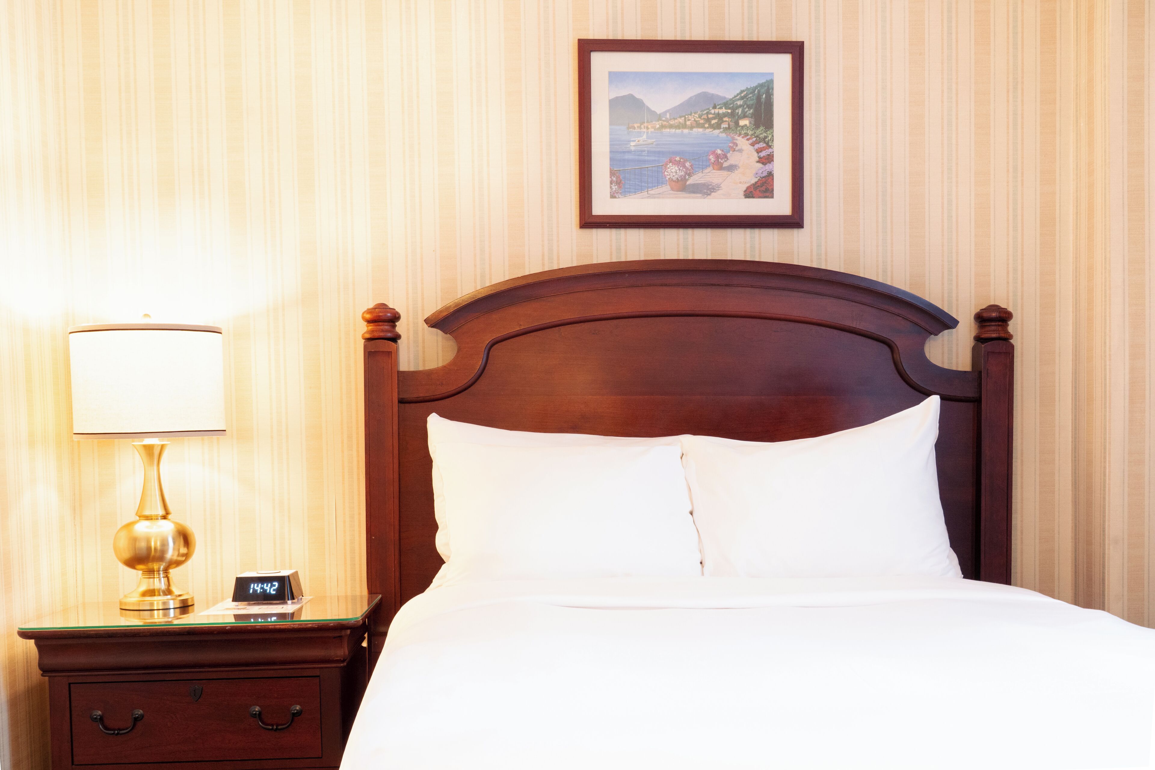 A neatly made bed with white linens, a wooden headboard, a bedside table with a lamp, and a framed picture on the wall.