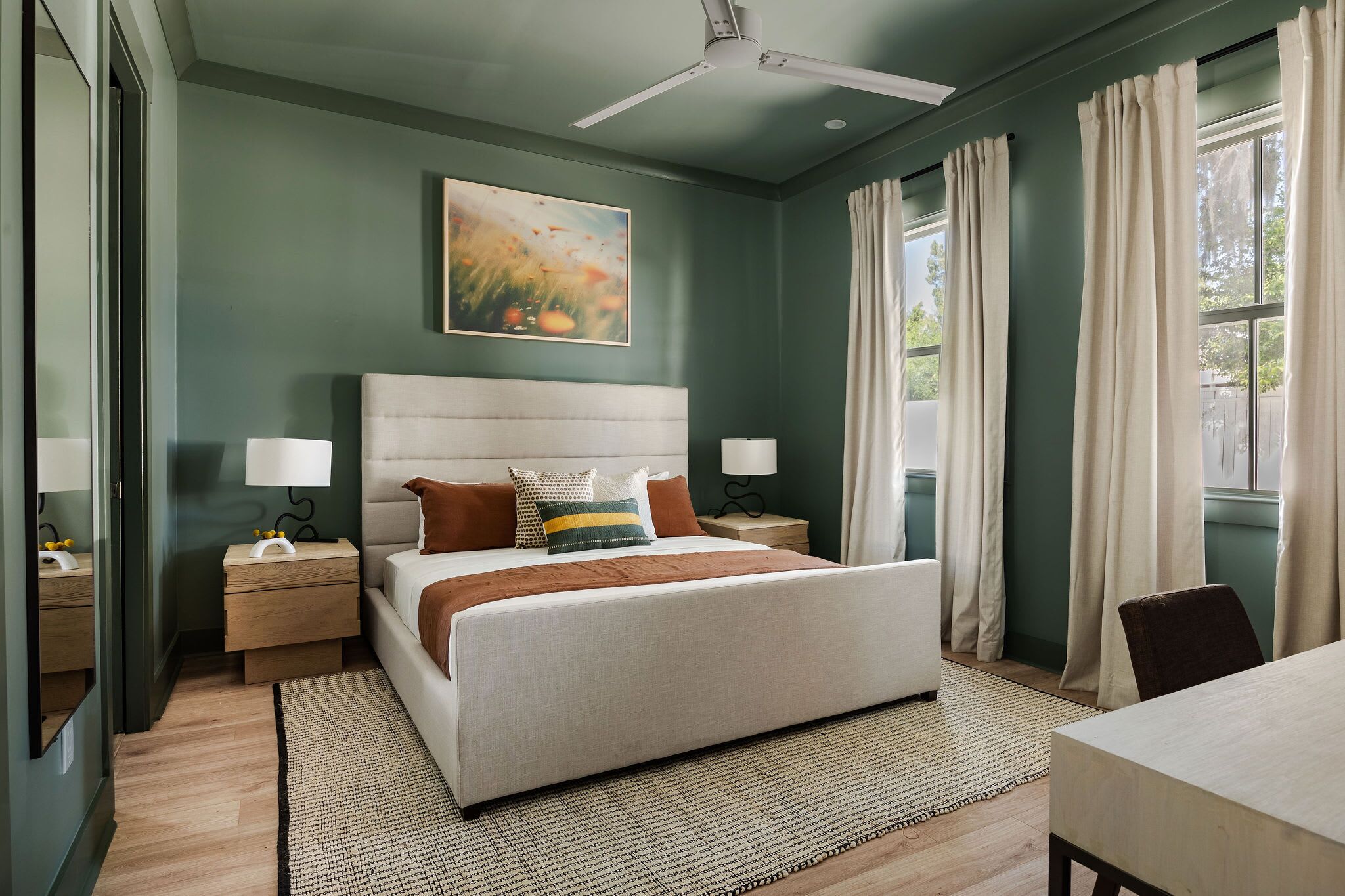 A bedroom with a large bed, two bedside tables with lamps, a ceiling fan, a painting on the wall, and a window with curtains.