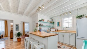 Une cuisine moderne avec des meubles blancs, un plan de travail en bois et des tabourets de bar. On y trouve des étagères avec des plantes et des ustensiles de cuisine, ainsi qu’une fenêtre offrant une belle vue.