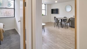 Habitación moderna y luminosa con comedor que incluye una mesa redonda y sillas, un televisor montado en la pared y una ventana con persianas.