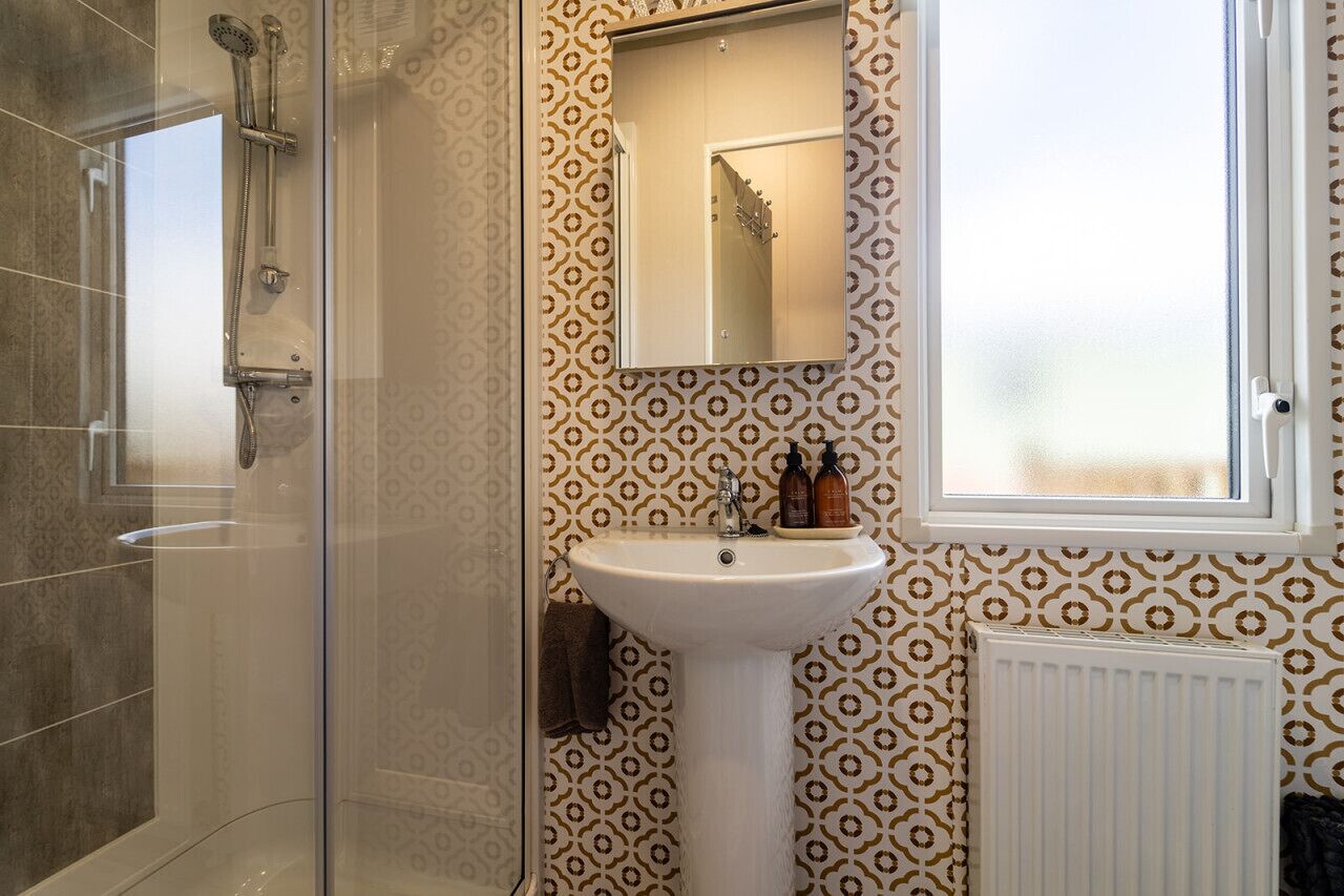 Une salle de bain avec une douche à parois de verre, un lavabo sur pied blanc et un papier peint à motifs.
