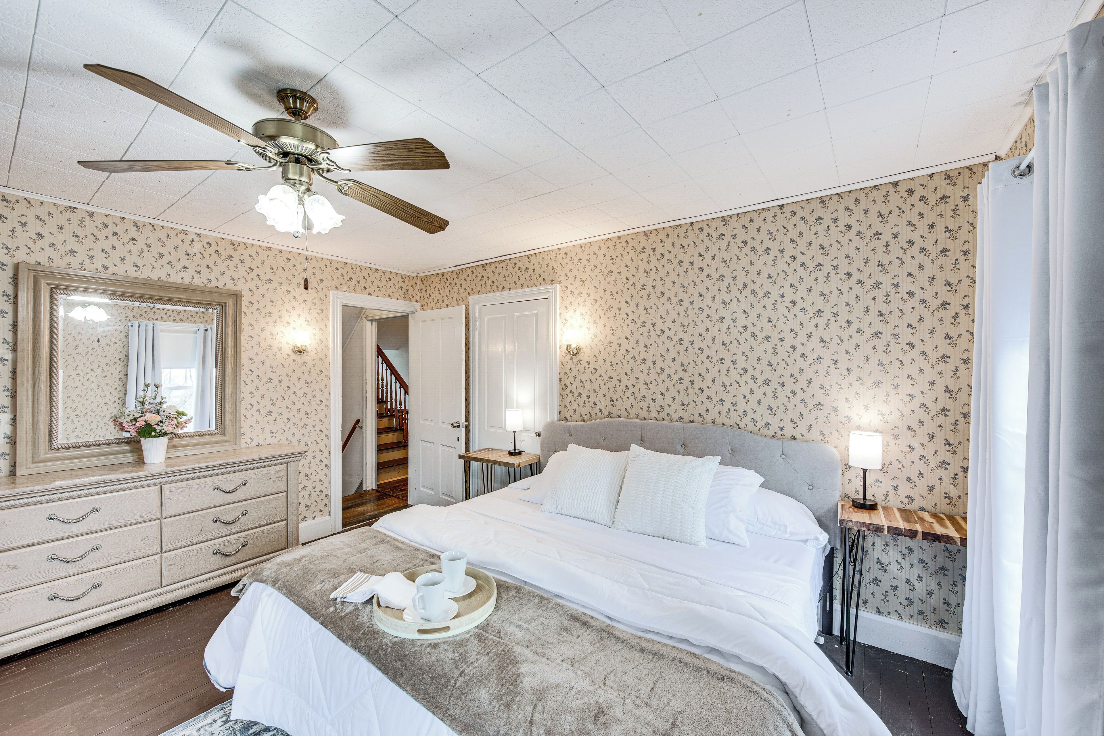 Een slaapkamer met een bed, een dressoir, een spiegel, een plafondventilator en een nachtkastje met een lamp.