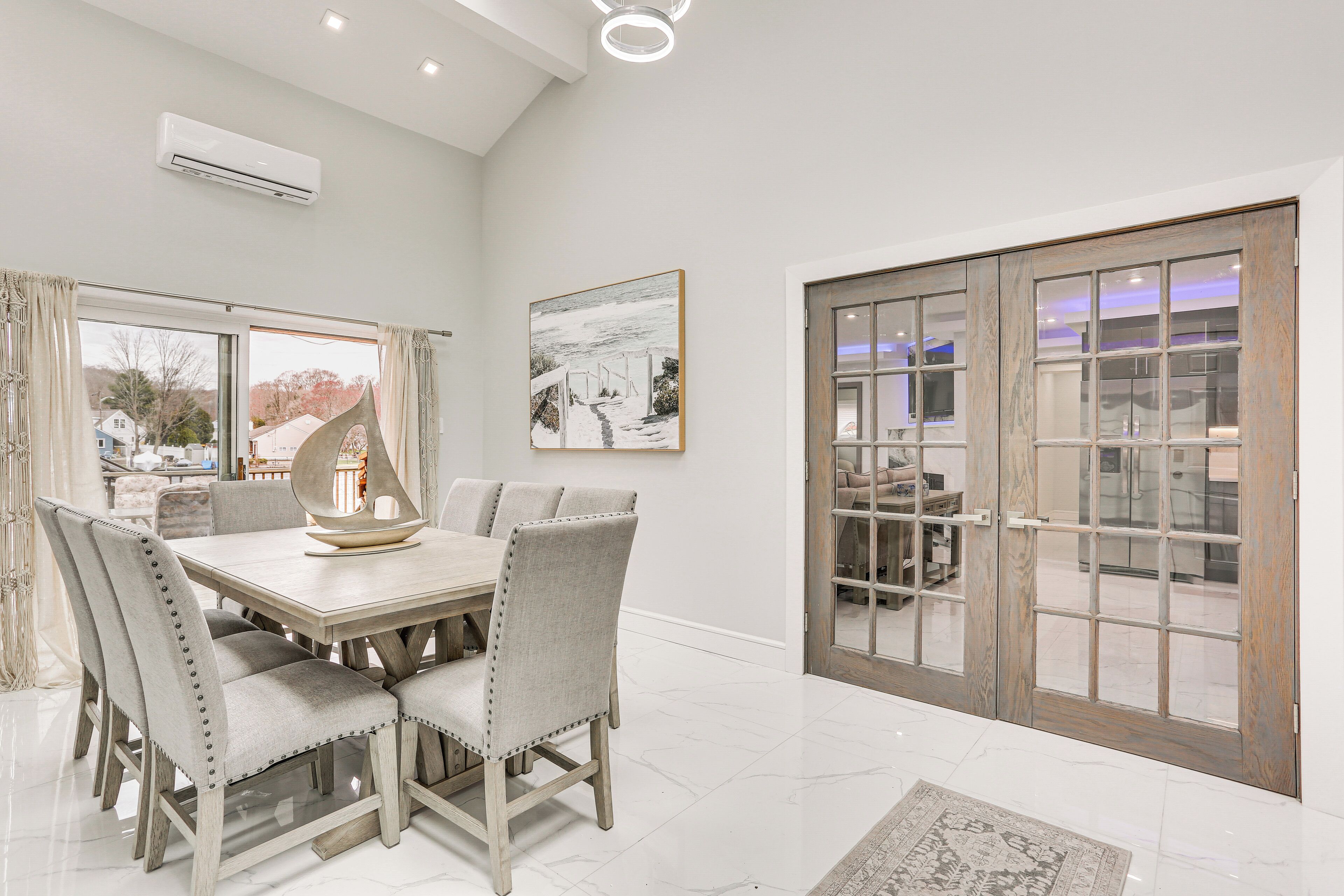 A dining area with a wooden table and upholstered chairs, a large window with curtains, and a sliding glass door leading to another room.