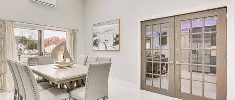 A dining area with a wooden table and upholstered chairs, a large window with curtains, and a sliding glass door leading to another room.