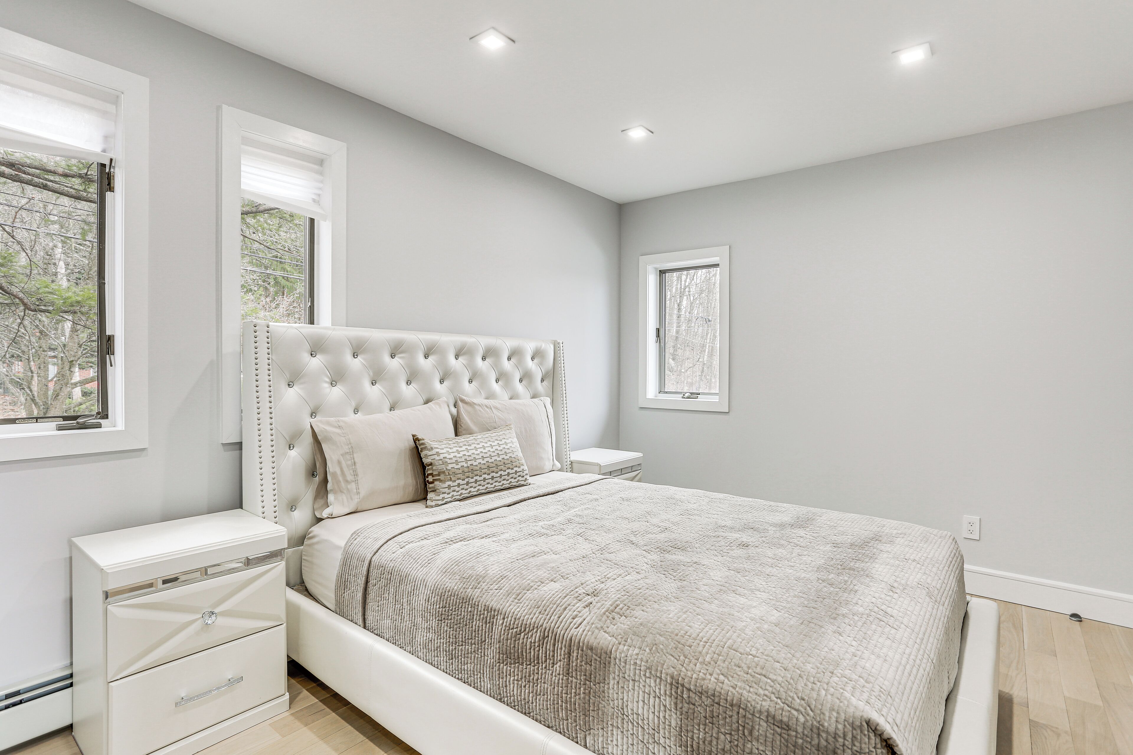 Une chambre à coucher avec une tête de lit capitonnée, un lit recouvert d’une couverture texturée, une table de chevet avec des tiroirs et une fenêtre équipée de stores.