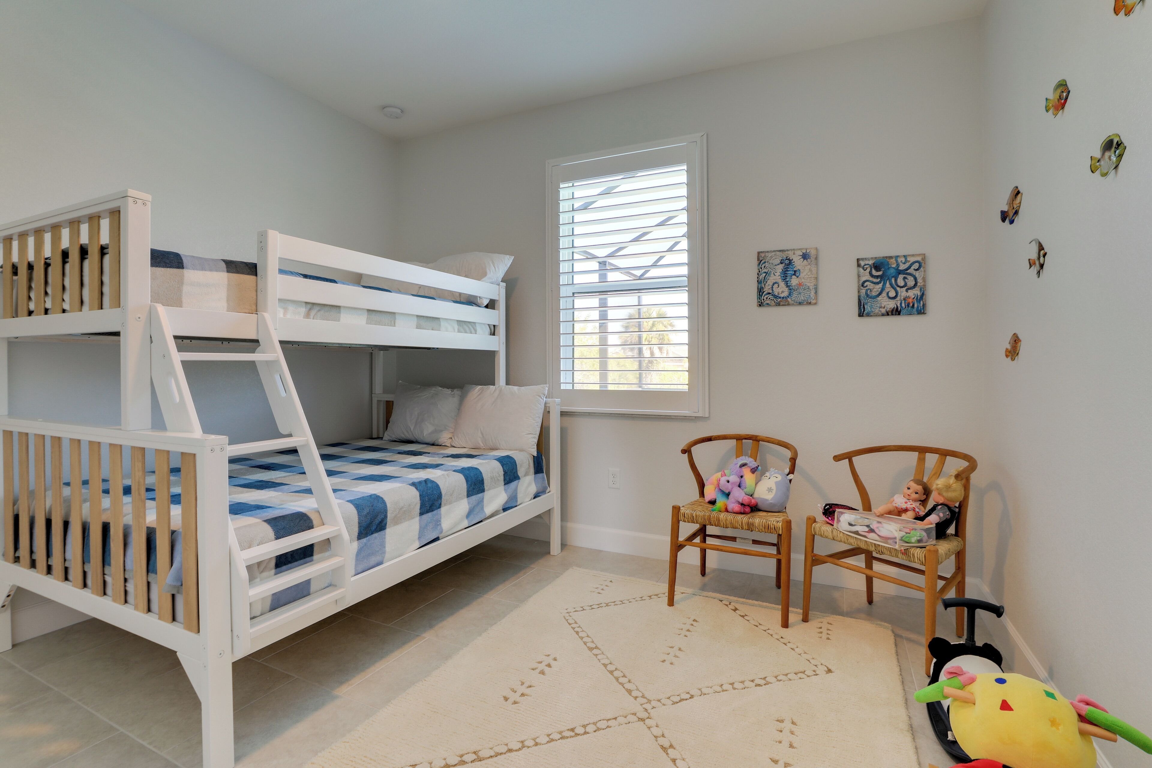 Une chambre d’enfant avec des lits superposés, une fenêtre avec des stores, deux chaises et des décorations murales.