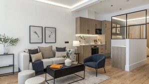 Una sala de estar moderna con sofá, mesa de centro y otomanas. Se observa una zona de cocina con armarios de madera y encimera.