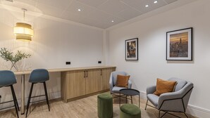 Un área de descanso moderna con una barra de madera, dos taburetes altos, un sillón gris con un cojín marrón y una mesa redonda.