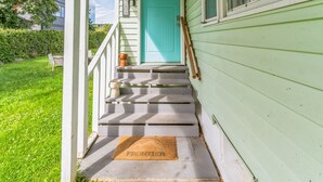 Une porte turquoise, ornée d’une boîte aux lettres et d’un paillasson sur lequel est inscrit le mot « FRONTIÈRE », mène à un porche doté de marches.