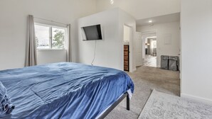 Quarto com cama, TV fixada na parede, janela com cortinas e piso coberto por carpete.