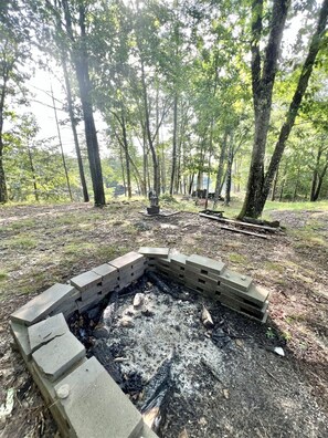 A circular stone fire pit surrounded by a forested area with trees and a bench in the background.