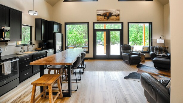 A spacious living area with a large wooden table, black leather sofas, and a black leather couch. There are three large windows providing natural light and a view of greenery outside. - Great Access just Off Hwy, Hot Tub, Open Plan w/ Parking! Close to Town! (Lead)