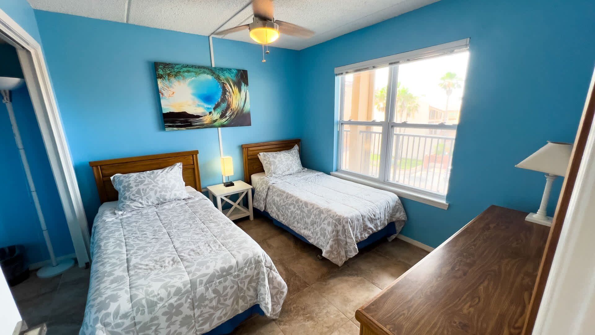 A bedroom with two beds, a ceiling fan, a painting, and a window with a view of palm trees.
