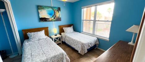 A bedroom with two beds, a ceiling fan, a painting, and a window with a view of palm trees.