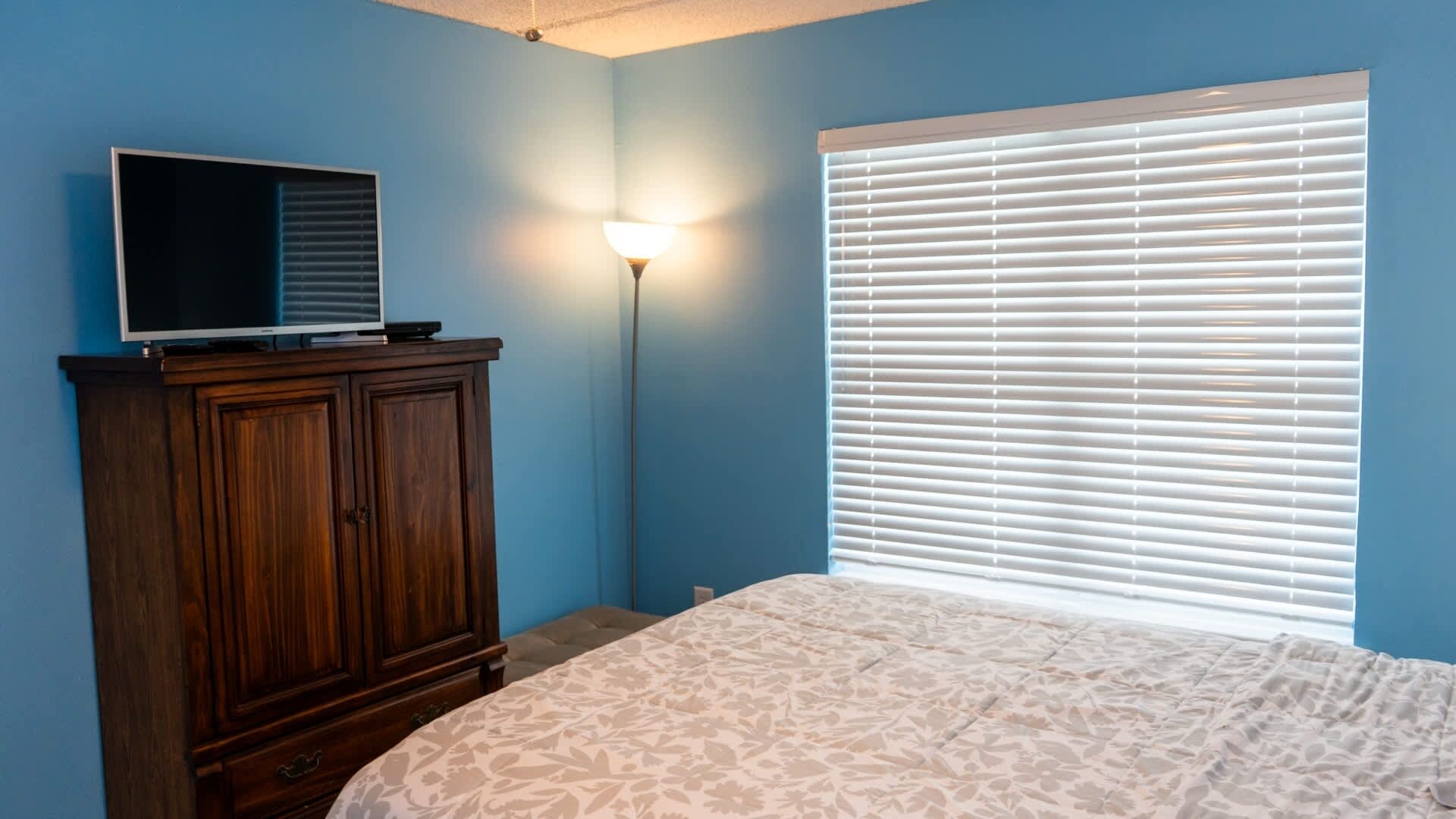Un dormitorio con cama, cómoda de madera, televisor encima y ventana con persianas.