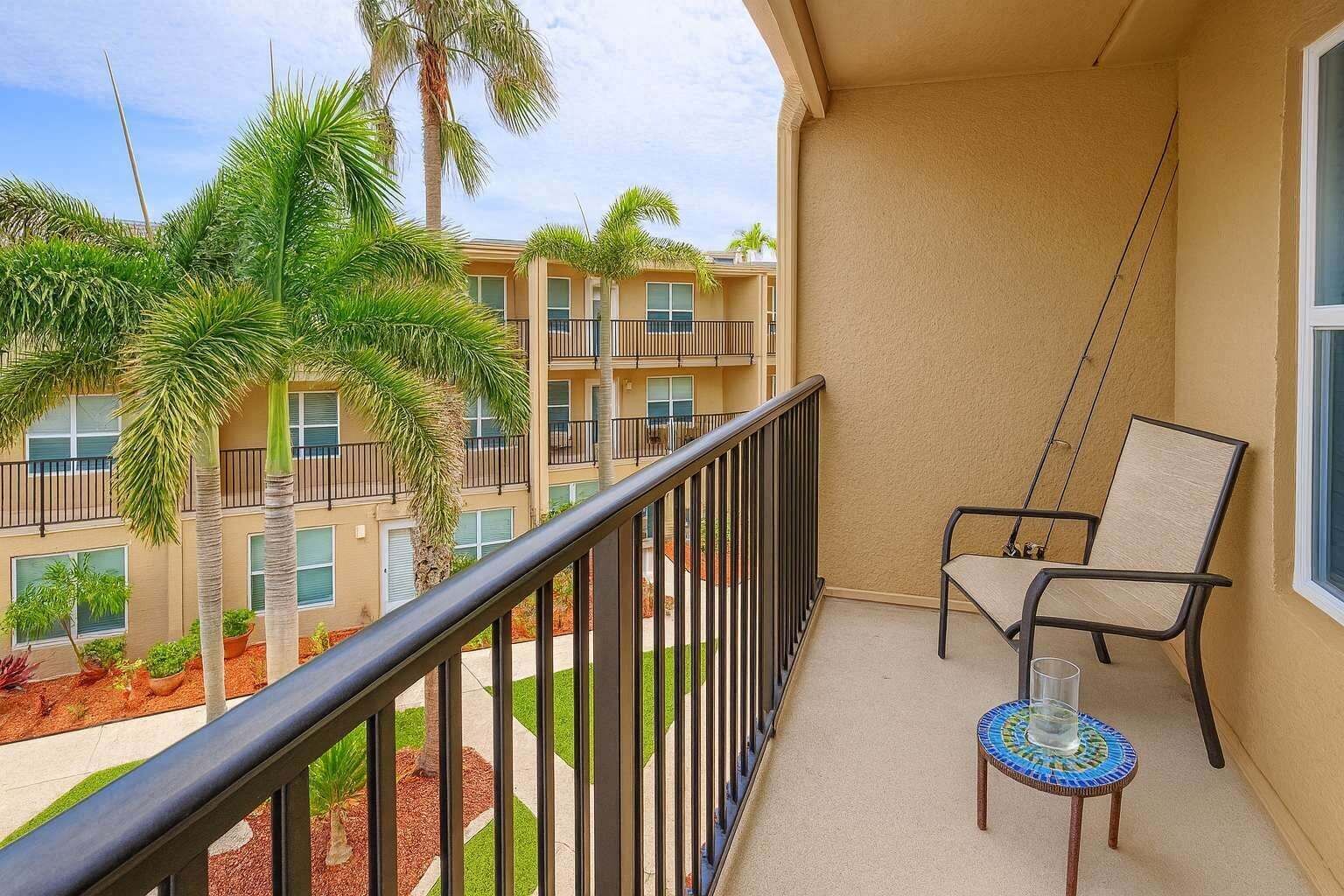A balcony with a chair and a small table, overlooking a building with multiple balconies and palm trees.