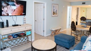 A living room with a TV, a small table, a blue couch, and a dining area with chairs. - Surfside Ii #109 2 Bedroom Home by RedAwning (South Padre Island)