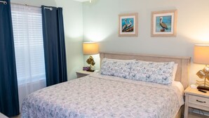 A bedroom with a bed, two bedside tables, a lamp, a curtain, and two framed pictures on the wall. - Surfside Ii #109 2 Bedroom Home by RedAwning (South Padre Island)