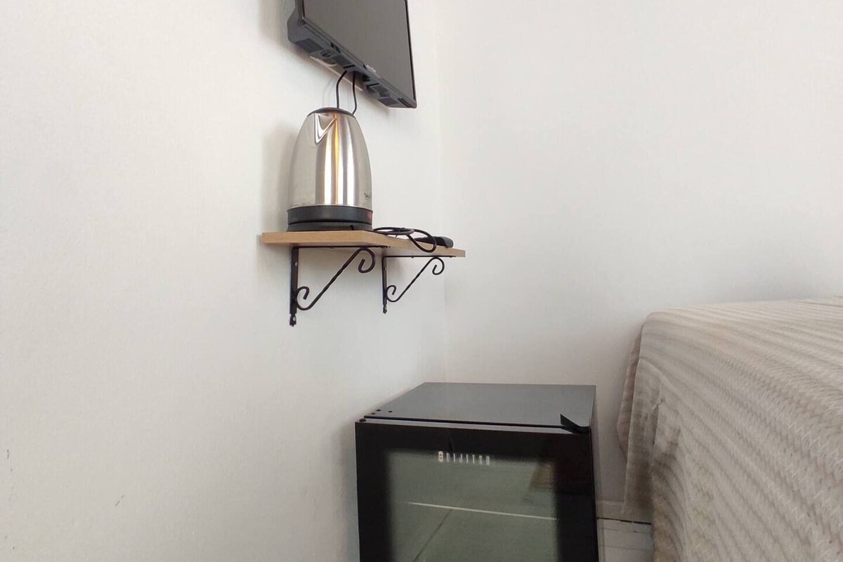 A small room with a television mounted on the wall, a small refrigerator, and a bed with a striped blanket.