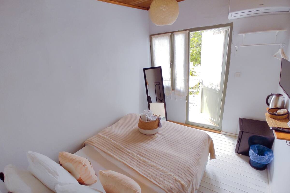 A bedroom with a bed, pillows, a bedside table with a kettle, a trash bin, and a window with curtains.