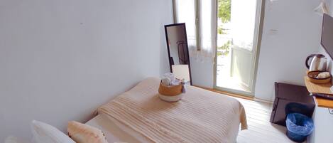 A bedroom with a bed, pillows, a bedside table with a kettle, a trash bin, and a window with curtains.
