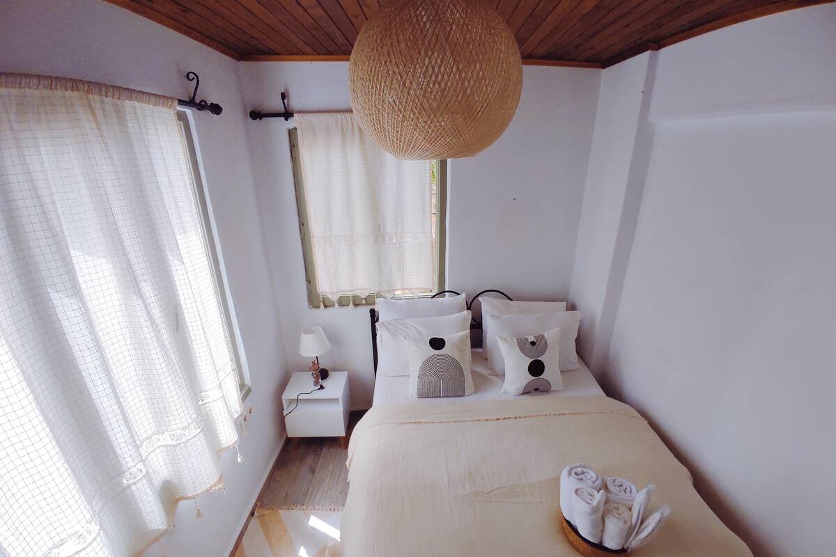 A bedroom with a bed, a bedside table, a lamp, a window with curtains, and a hanging light fixture.