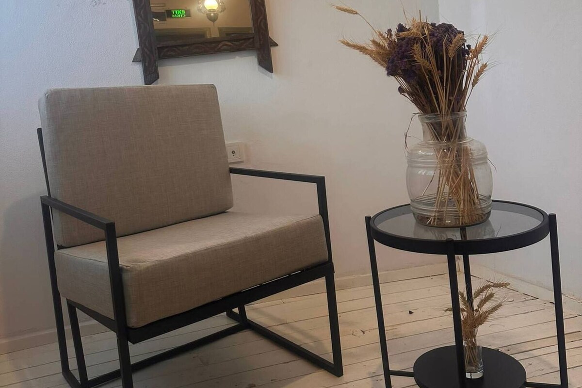A modern room with a minimalist chair, a small table with a vase of dried plants, and a wall-mounted mirror with a light fixture.