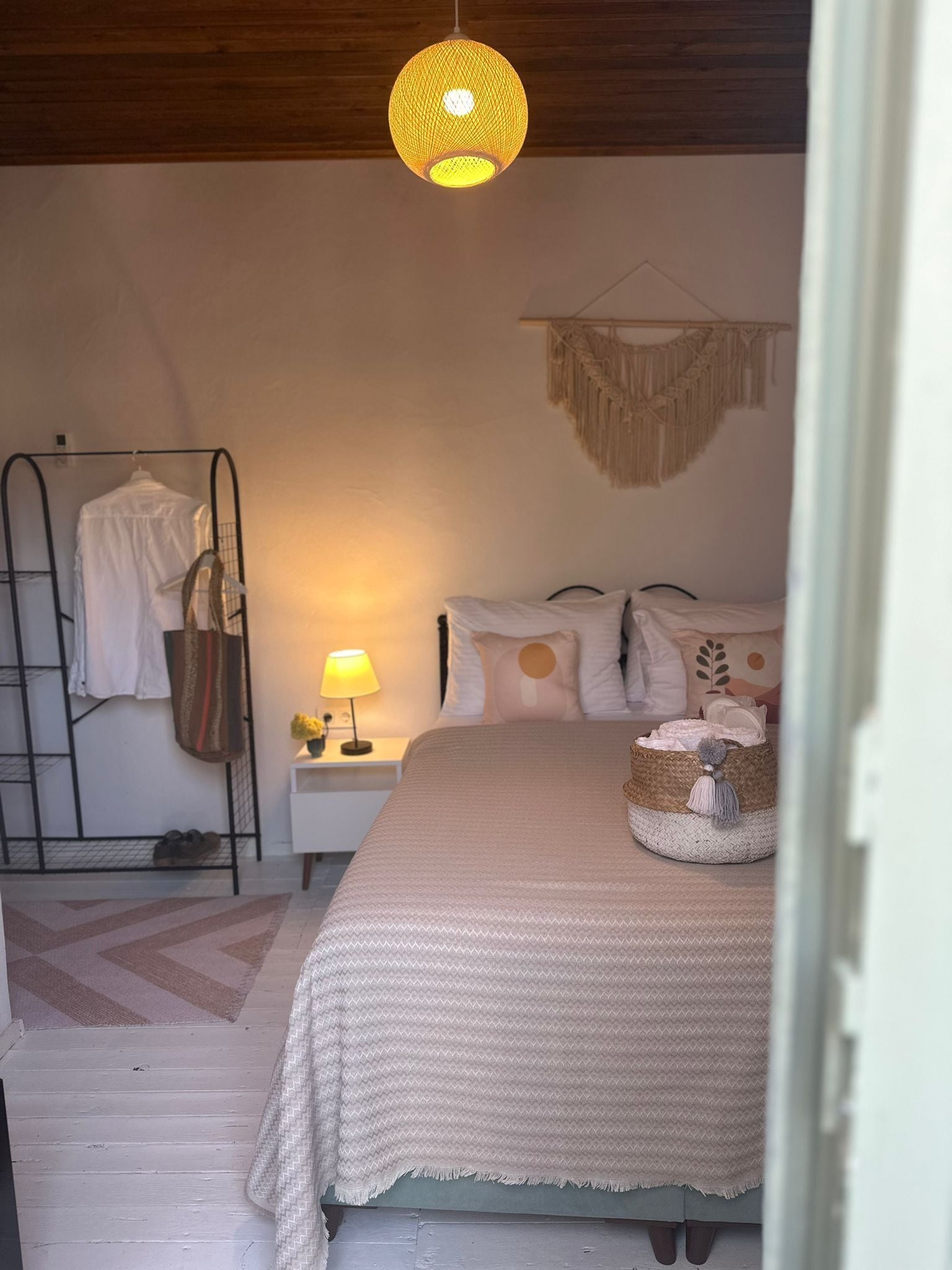 A cozy bedroom with a bed, a hanging macrame wall decoration, a bedside lamp, and a clothing rack.