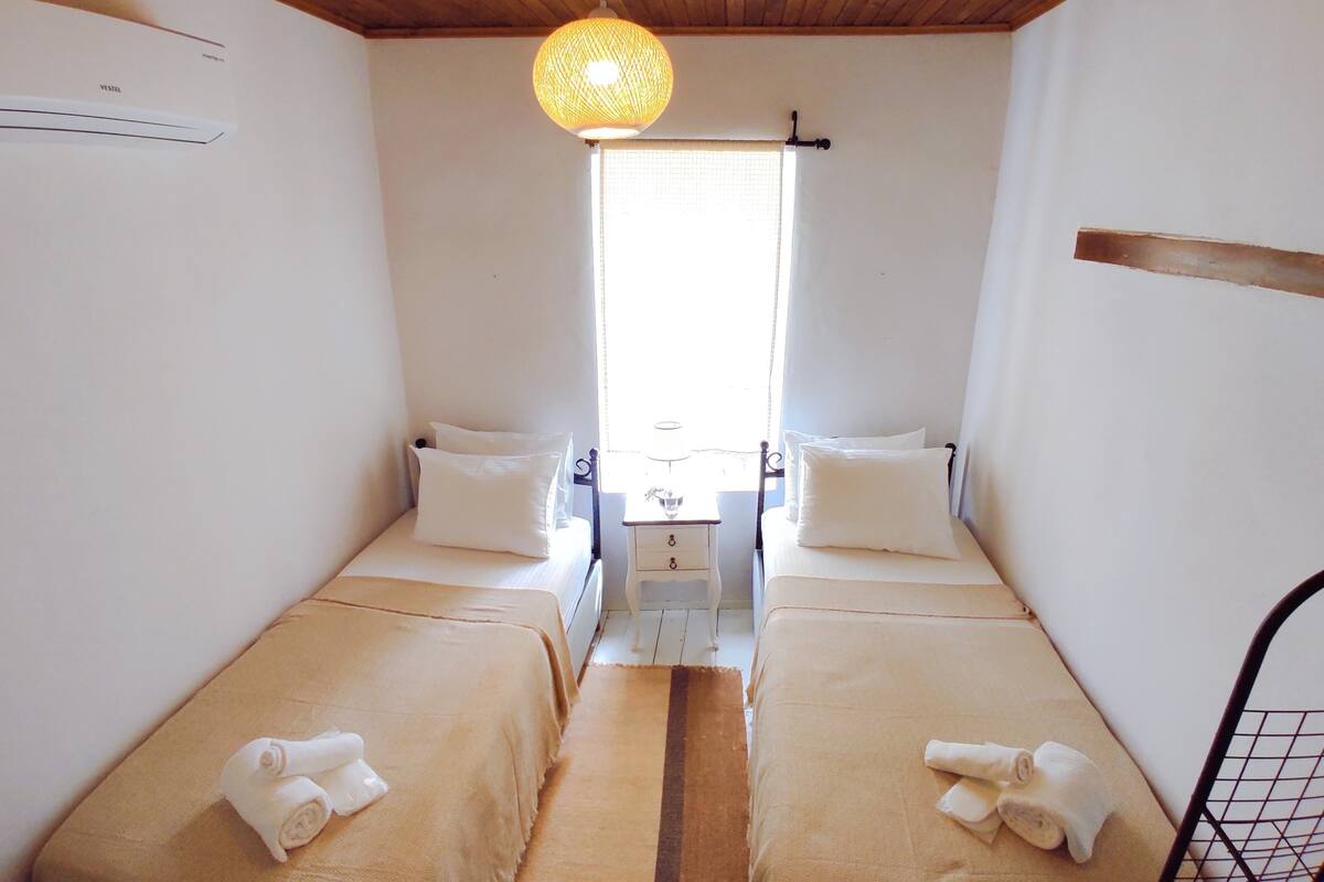 A small, clean room with two single beds, a wooden ceiling, a hanging light fixture, and a window with a curtain.