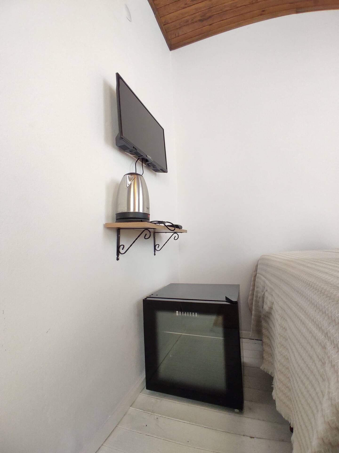 A small room with a television mounted on the wall, a small refrigerator, and a bed with a striped blanket.
