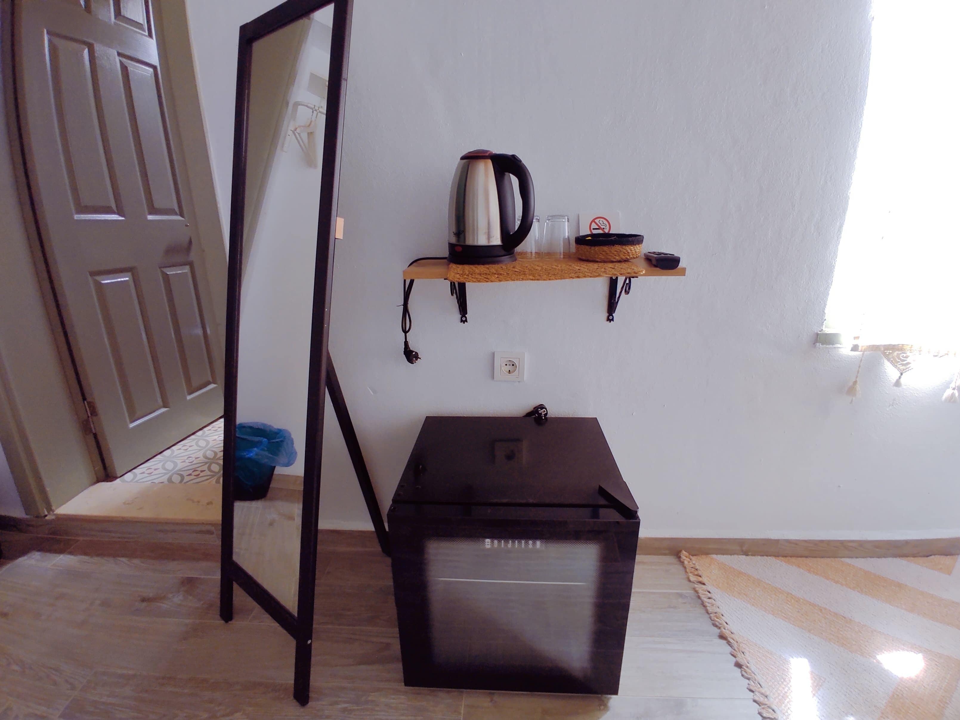 A room with a wooden floor, a black cabinet, a mirror leaning against the wall, and a shelf with a kettle and cups.