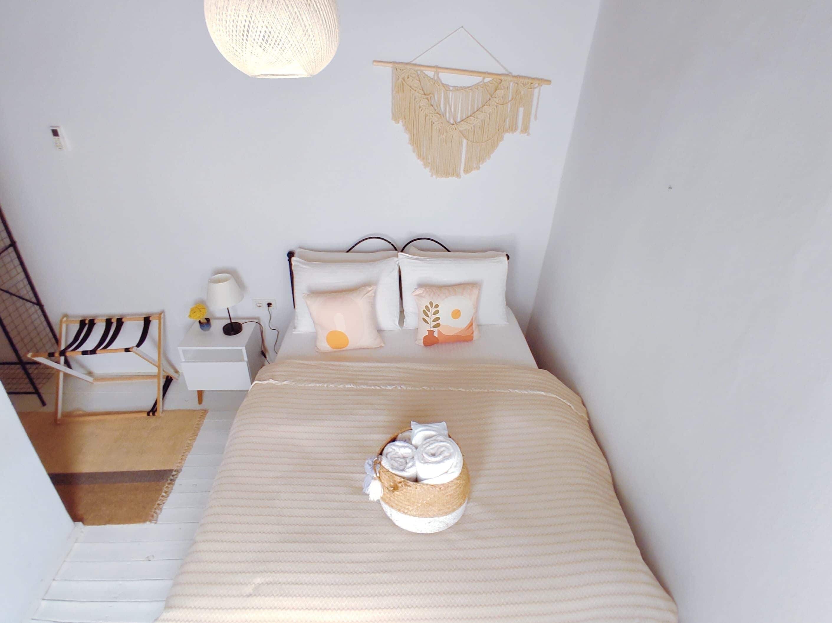 Une chambre bien rangée, avec un lit, une table de chevet, une lampe, une décoration en macramé suspendue et une petite chaise en bois.