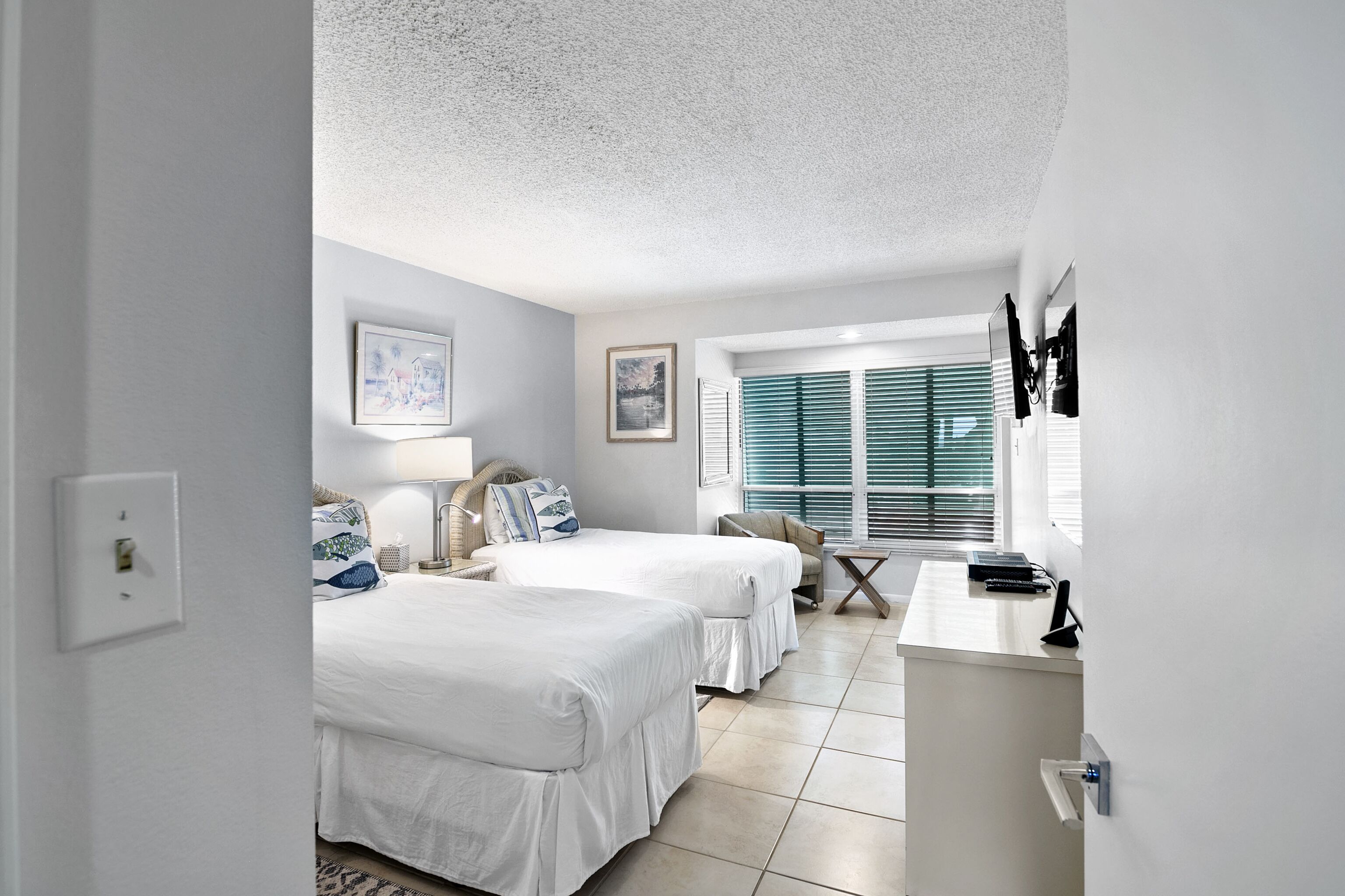 A hotel room with two beds, a desk, a chair, and a window with blinds.