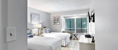 A hotel room with two beds, a desk, a chair, and a window with blinds.