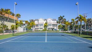 Una cancha de tenis con red, rodeada por una cerca, palmeras y casas al fondo.