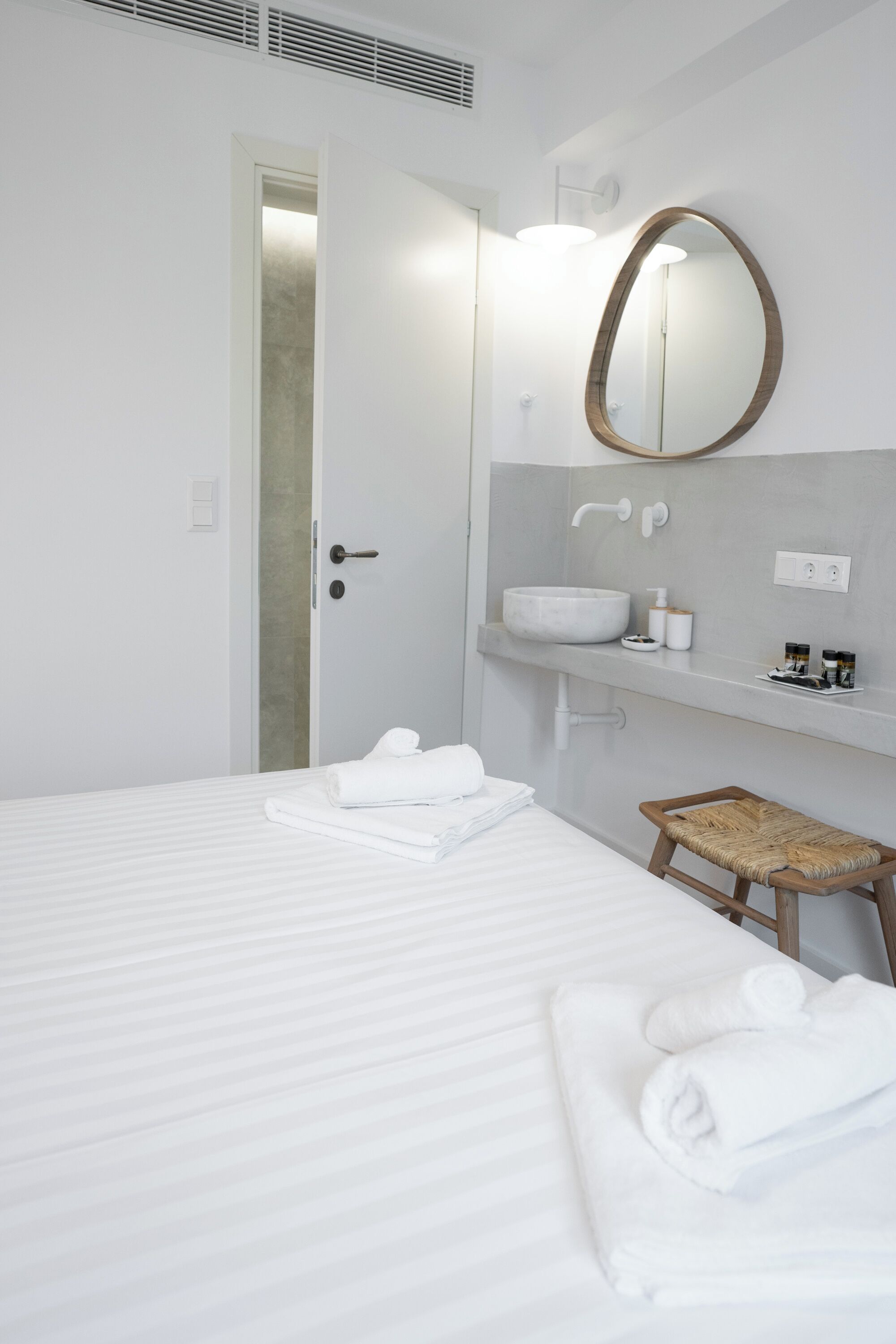 Ett hotellrum med en säng, en träpall, en stor oval spegel, ett vitt handfat och en dörr.
