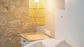 A bed with white linens and two pillows, a wooden nightstand with a lamp, and a stone wall.