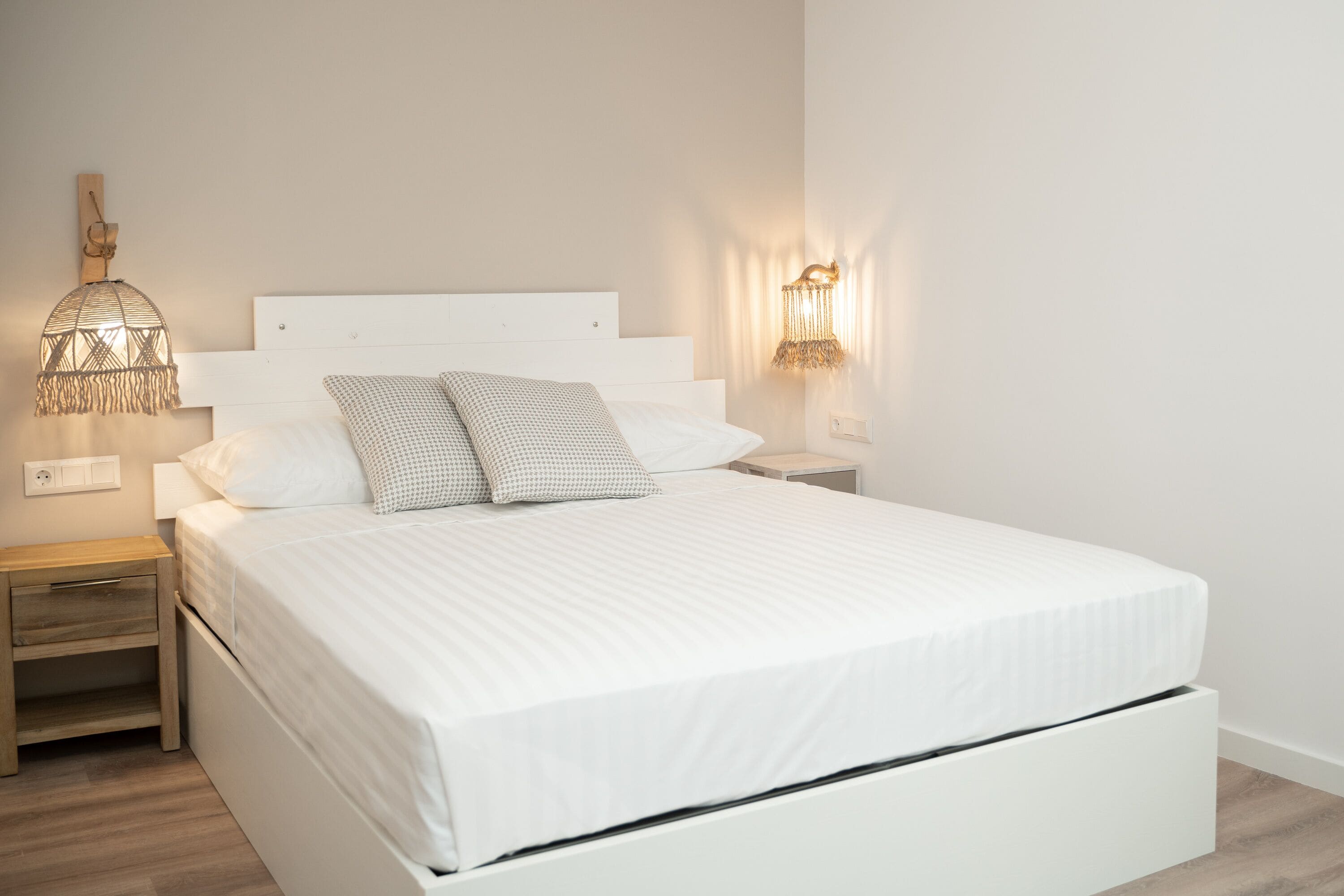 A neatly made bed with white linens and a wooden headboard, a bedside lamp, and a wall-mounted light fixture.