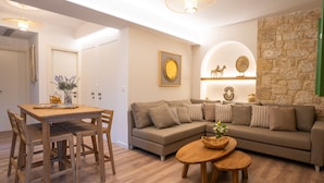 A modern living room with a beige sofa, wooden dining table, and chairs. There is a stone accent wall and a ceiling light fixture. - Olvio Suites (West Mani)