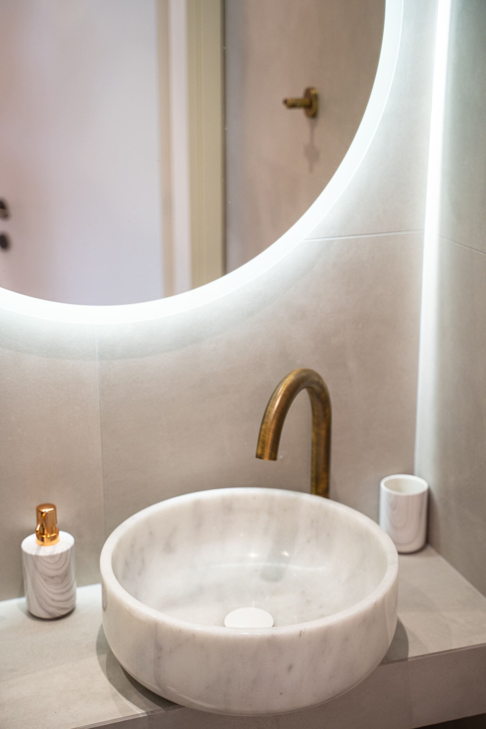 A bathroom with a round marble sink, a gold faucet, and a white marble cup on the counter.