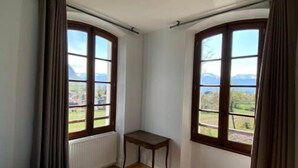 Una habitación con una cama, dos ventanas con cortinas, una mesita y vistas a montañas.