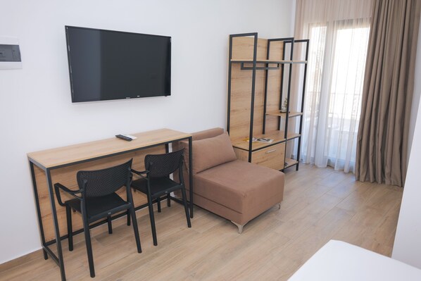 Una habitación moderna con un escritorio de madera, dos sillas negras, un sofá marrón, un televisor montado en la pared y una ventana con cortinas translúcidas.