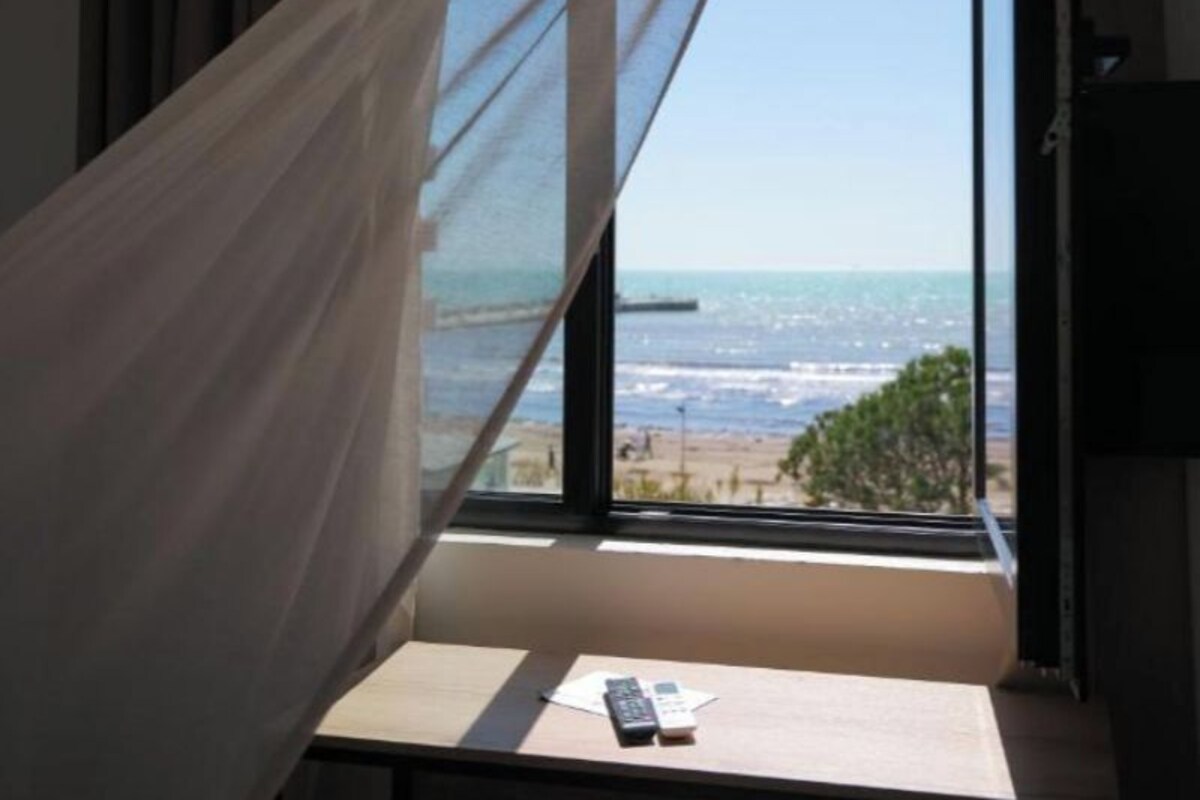 Una habitación con un ventanal grande, una cama con cortina translúcida, una mesita con un control remoto y vistas a la playa y al océano.