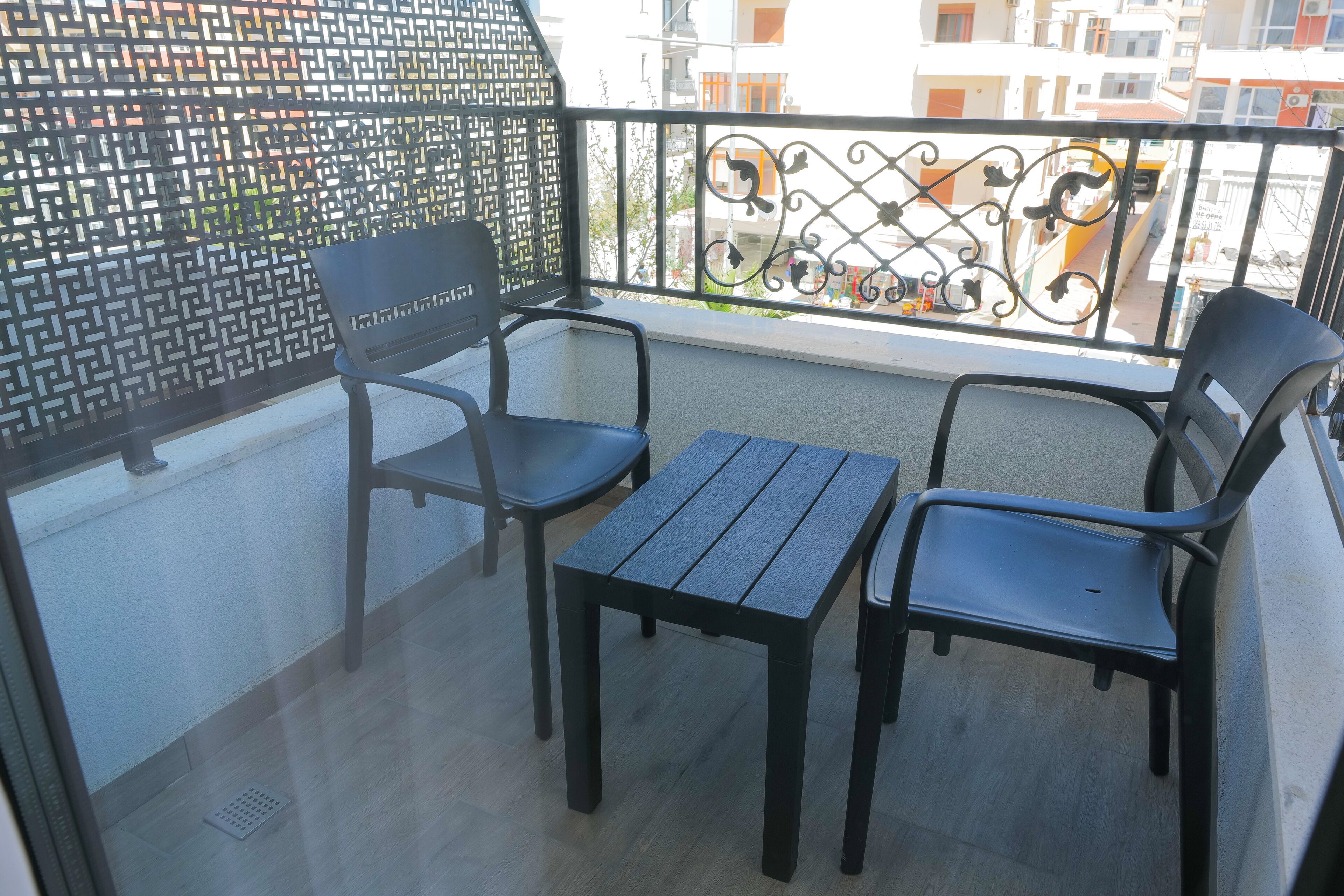 A balcony with two chairs and a table, a decorative metal railing, and a view of buildings in the background.